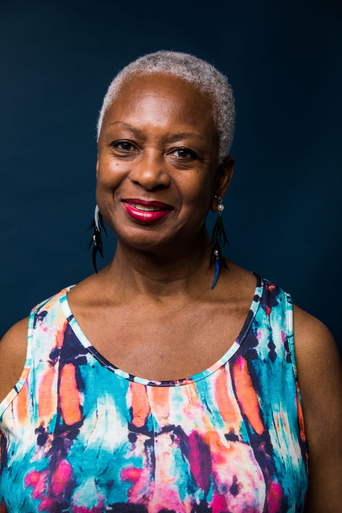 Woman with cropped gray hair wearing a multicolored dress and large dangling earrings gazes at the camera